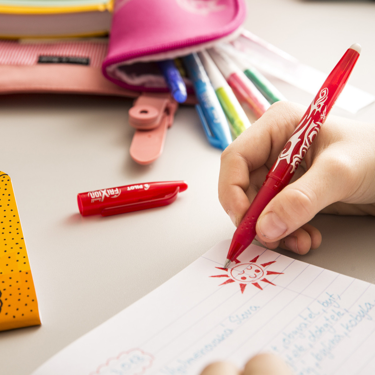 Child drawing a sun with PILOT’s FriXion Stick Erasable Gel Pen, surrounded by a colourful array of pens, ideal for kids' creative projects