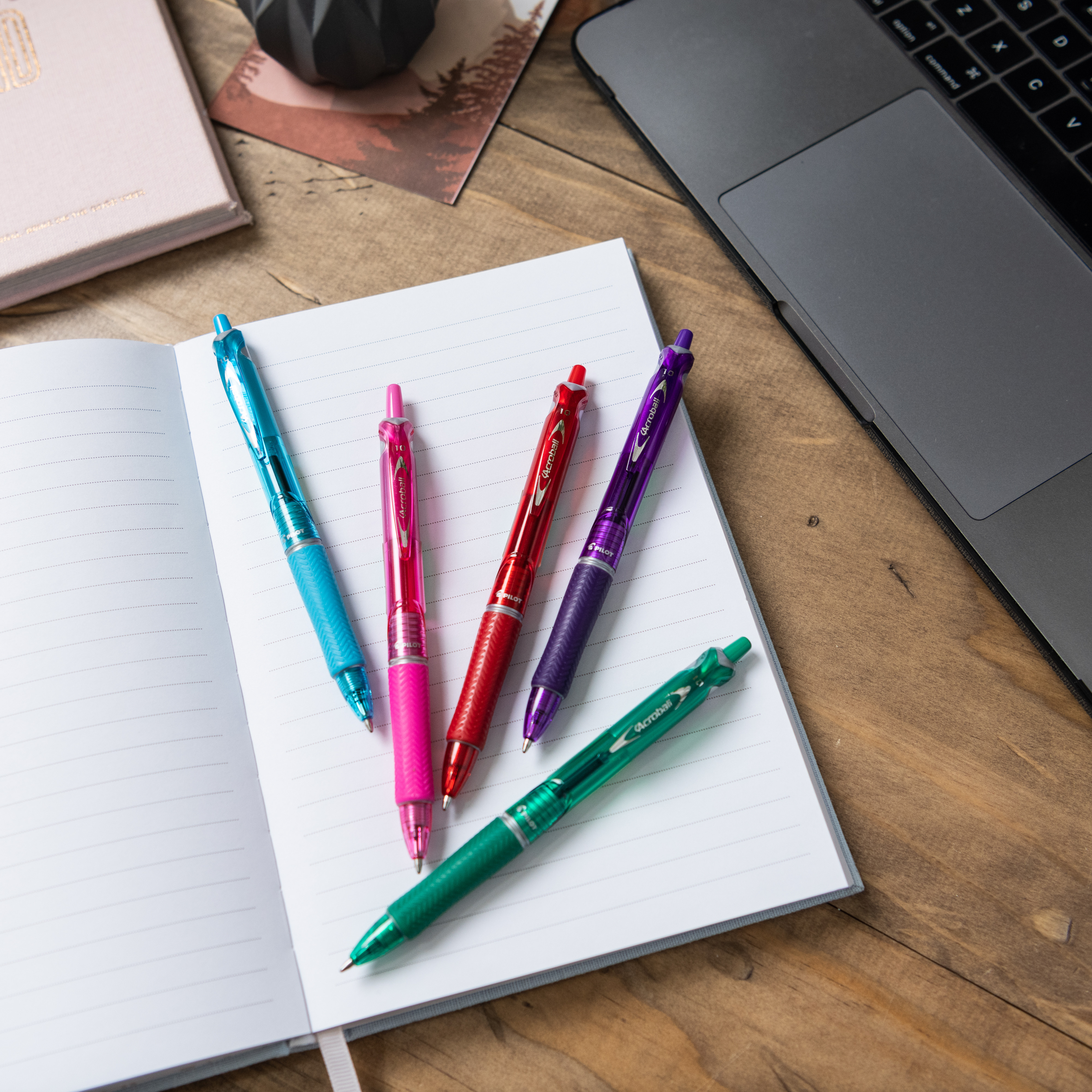 Set of colourful Pilot Acroball pens arranged on an open notebook near a laptop.