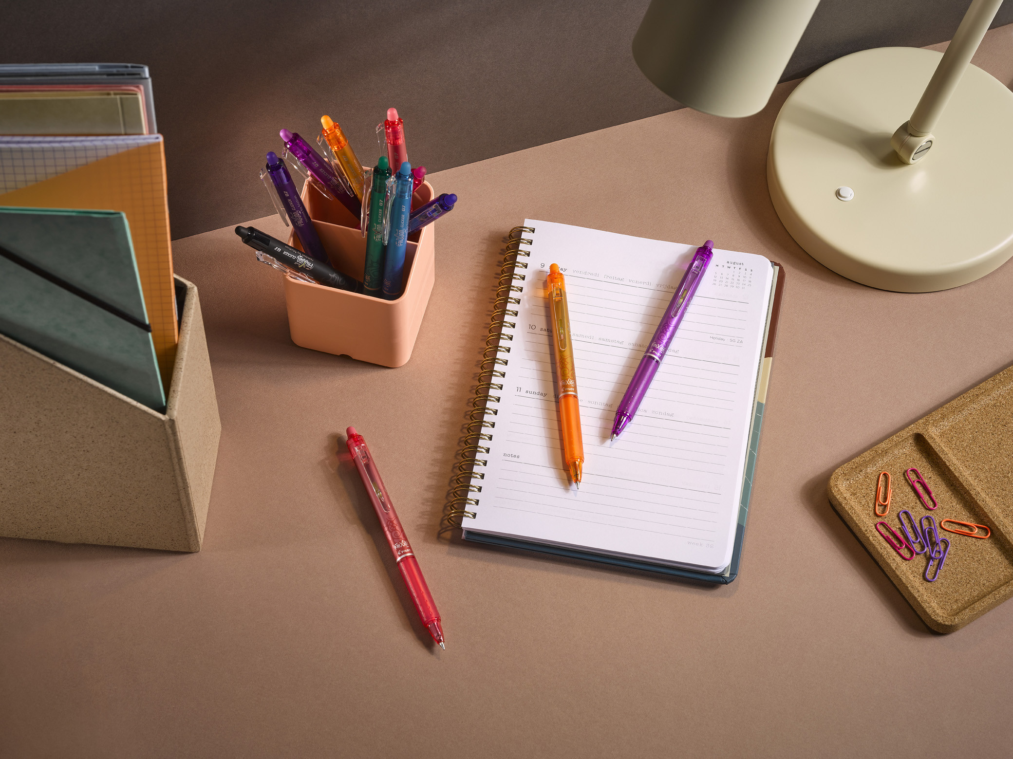 Assortment of Pilot FriXion Ball Clicker pens in various colours on a study desk with an open notebook.