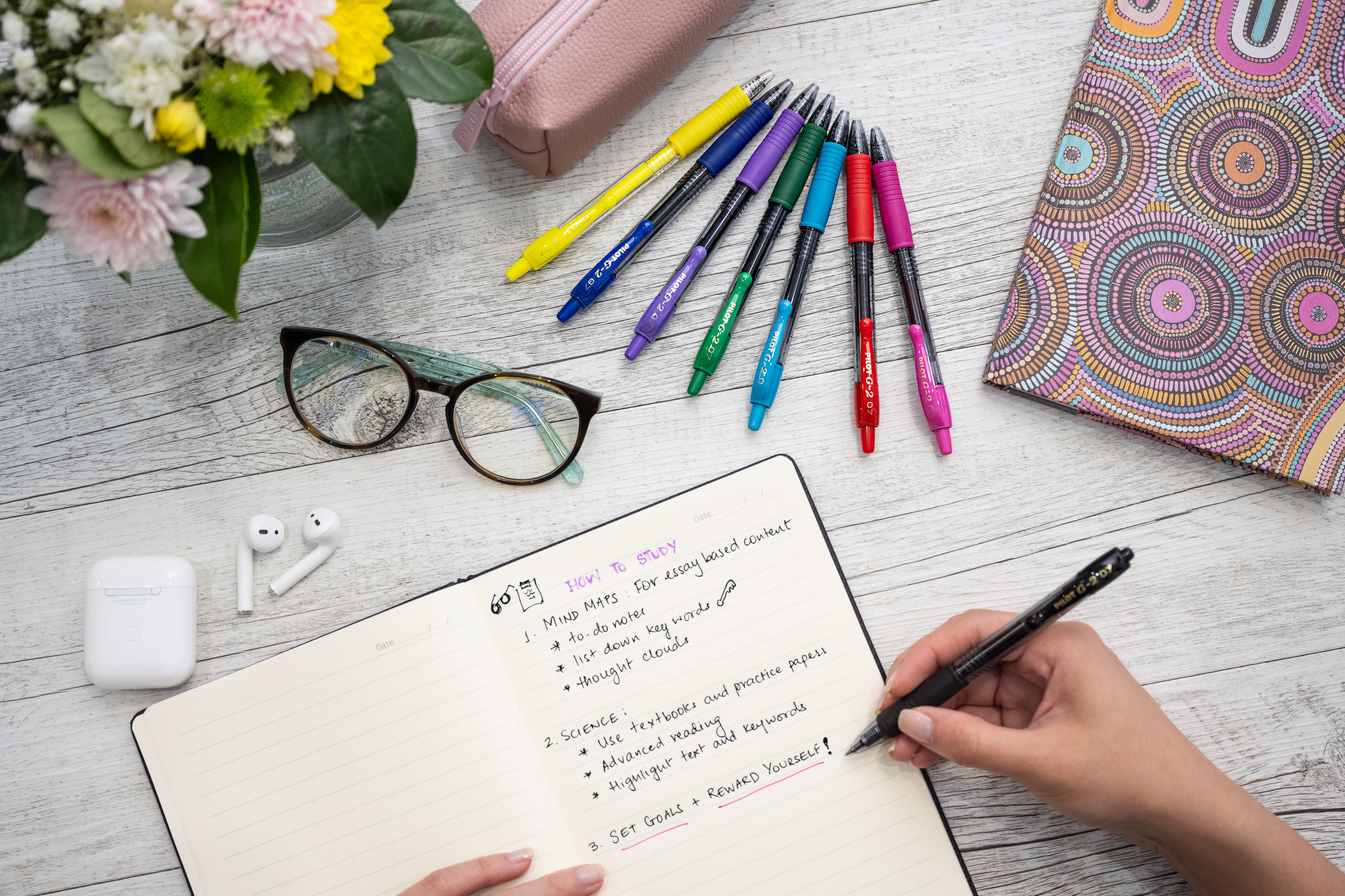 Person writing colourful study notes with G-2 Retractable Gel Pens, on a desk with glasses, pencil case and notebooks.