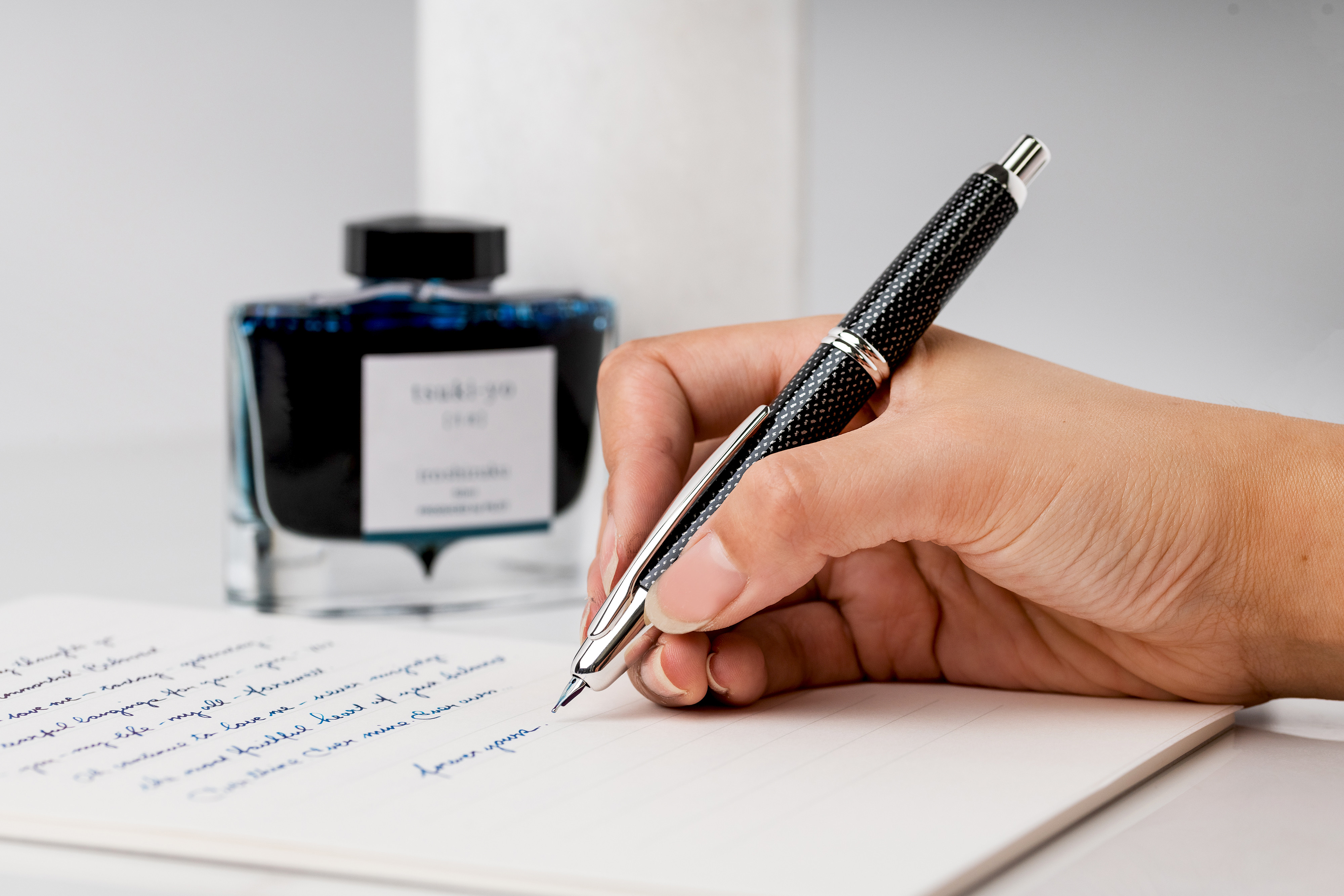 A hand writing with PILOT Capless Splash Retractable Fountain Pen with iroshizuku Fountain Pen Ink Bottle in the background, in the shade tsuki-yo.