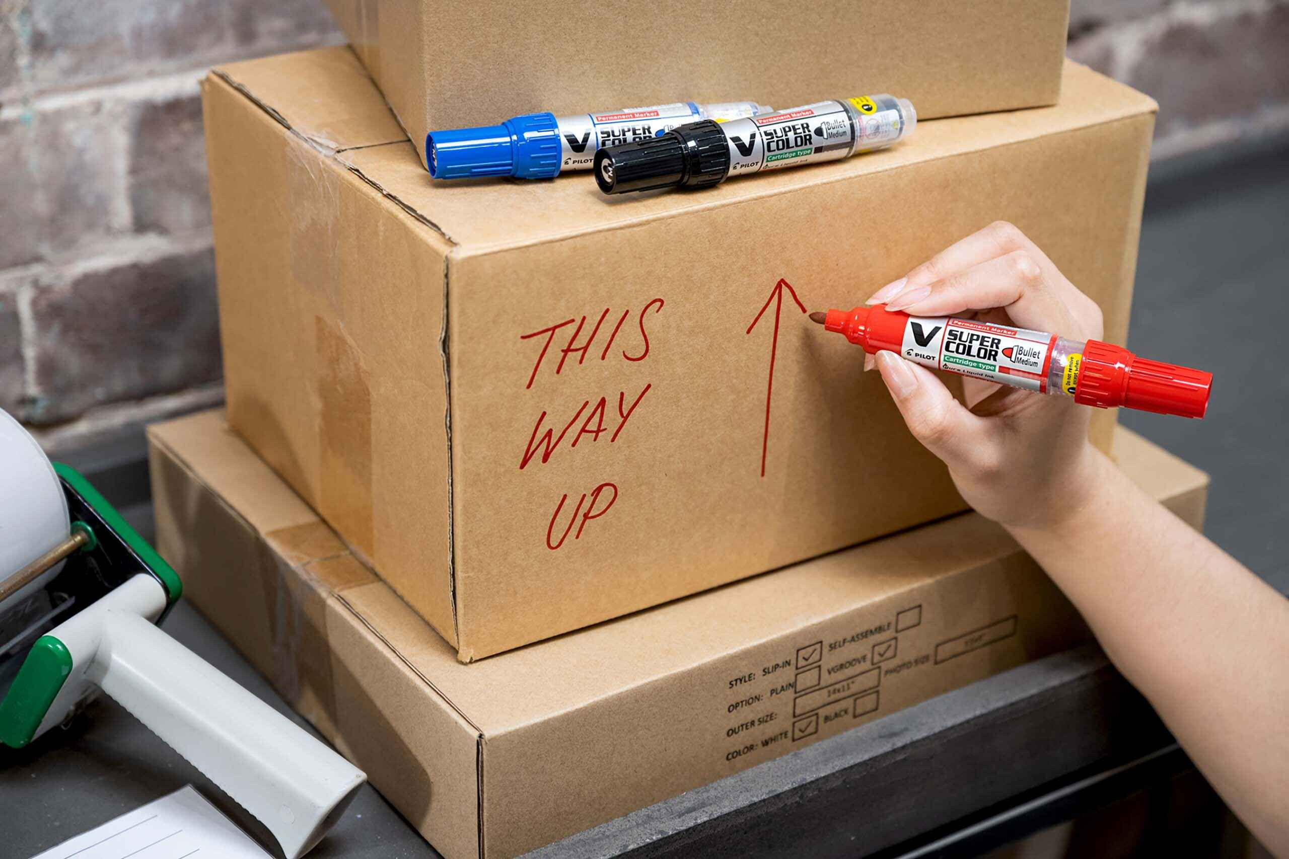 V Super Color Permanent Marker in red writing “This Way Up” on a cardboard box. 