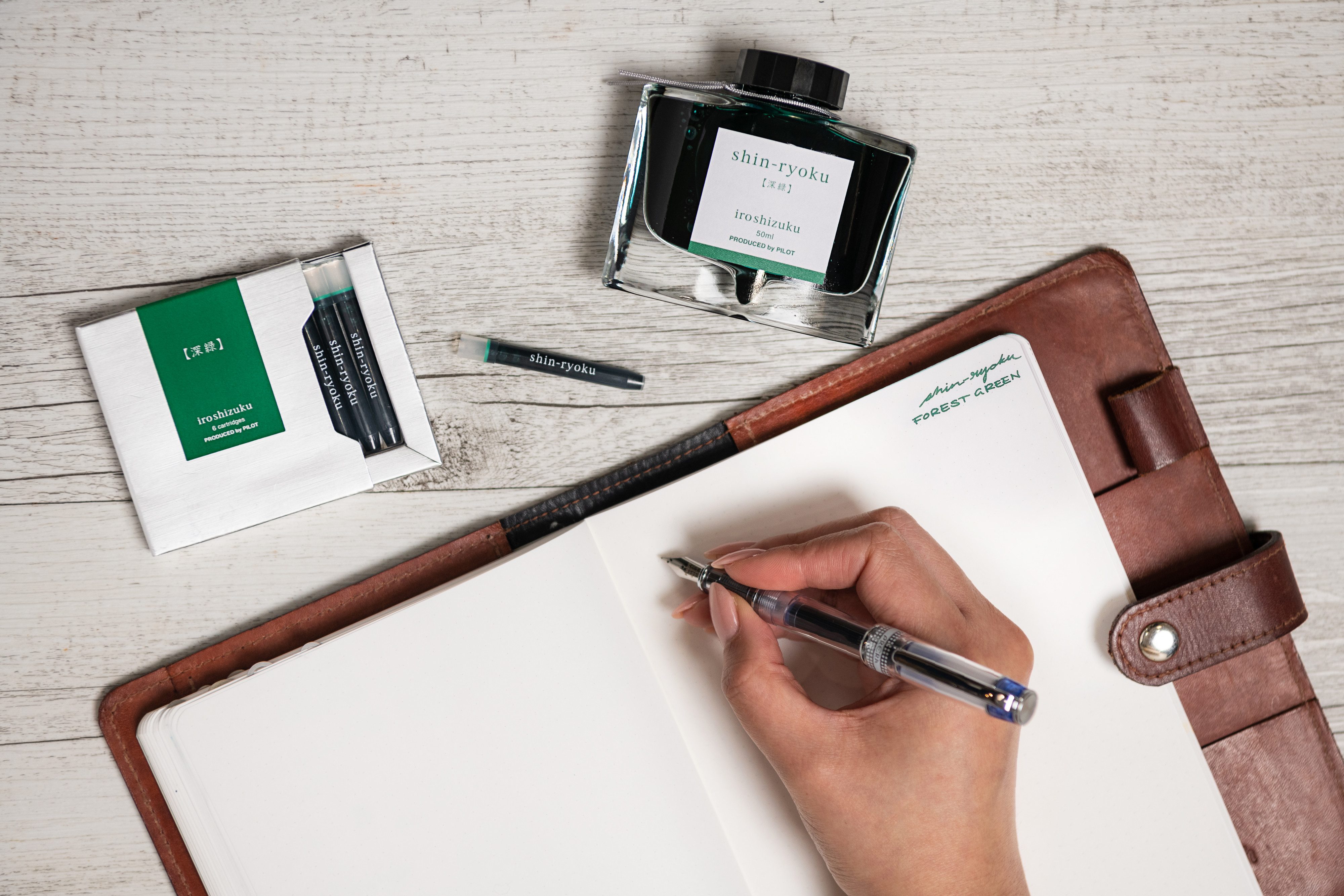Pilot Prera Transparent fountain pen, writing in green using Pilot iroshizuku ink in shin-ryoku, which translates to Forest Green in Japanese. The person is writing on a blank page in a leather covered notebook, with a glass bottle of the iroshizuku ink, as well as a pack of iroshizuku ink cartridges.