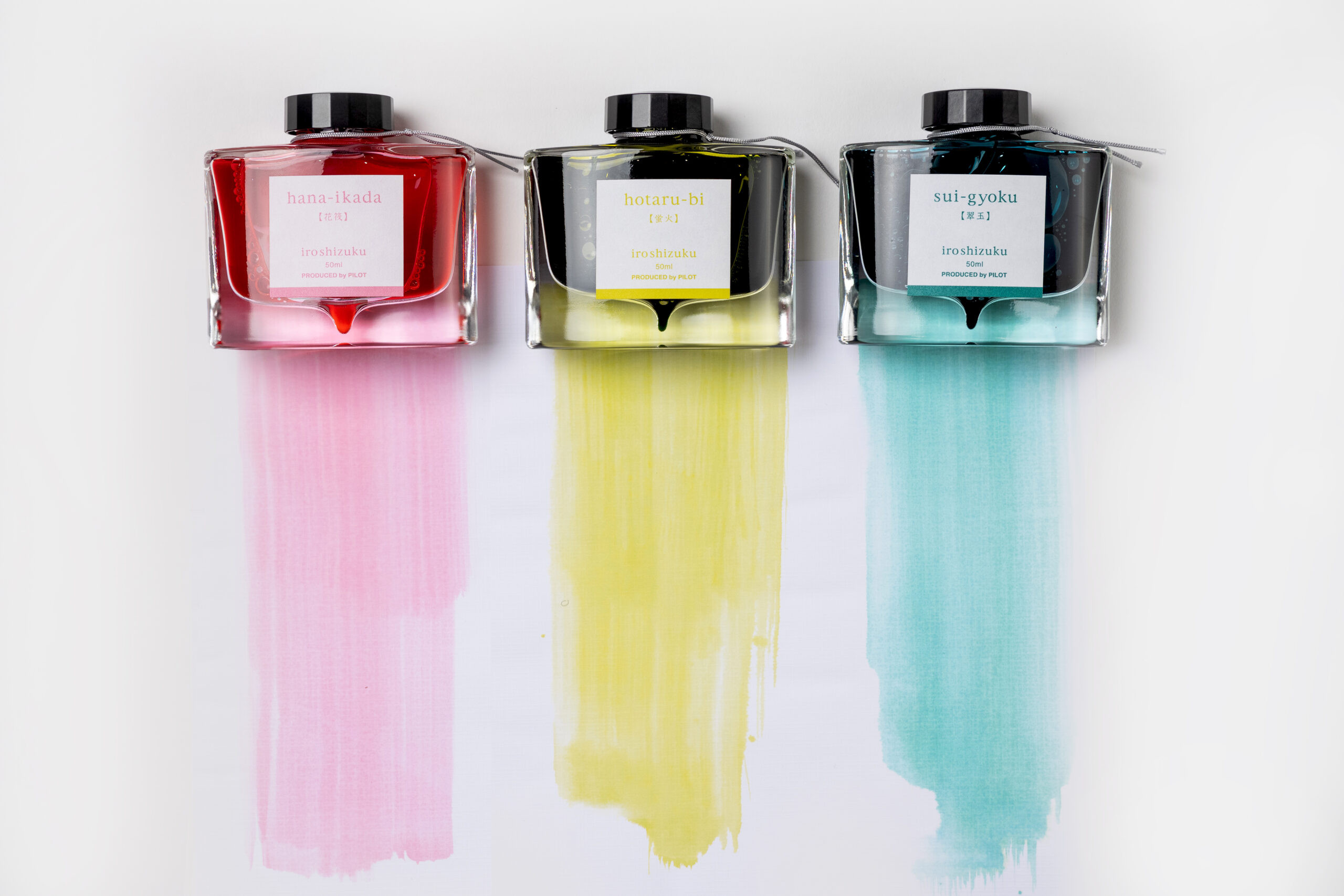 Three long swatches watch of Pilot iroshizuku ink, created using a broad paintbrush, sits under their corresponding glass bottles on a large piece of paper. The left bottle holds a pink ink, called hana-ikada, which translates to Cherry Blossom Petals in Japanese. The middle bottle is a yellow-green ink, called hotaru-bi, which translates to Firefly Glow. And the right bottle holds an ink called sui-gyoku, which translates to Emerald Green