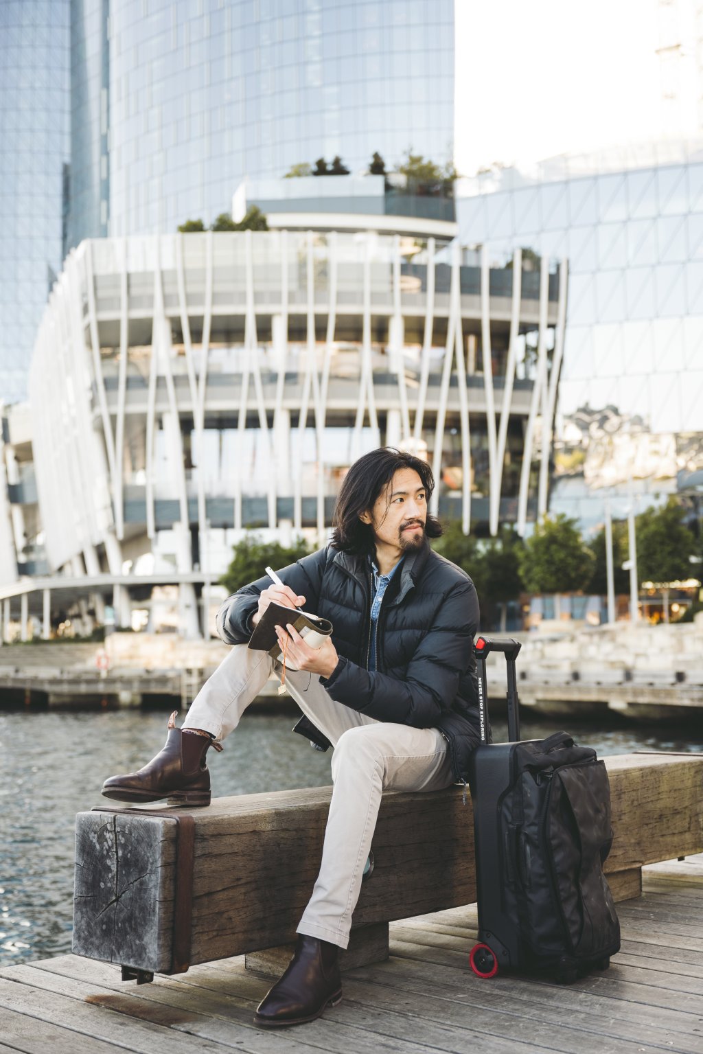 Man writing with pen and paper by the waterfront, taking an analogue pause from digital tools during travel.