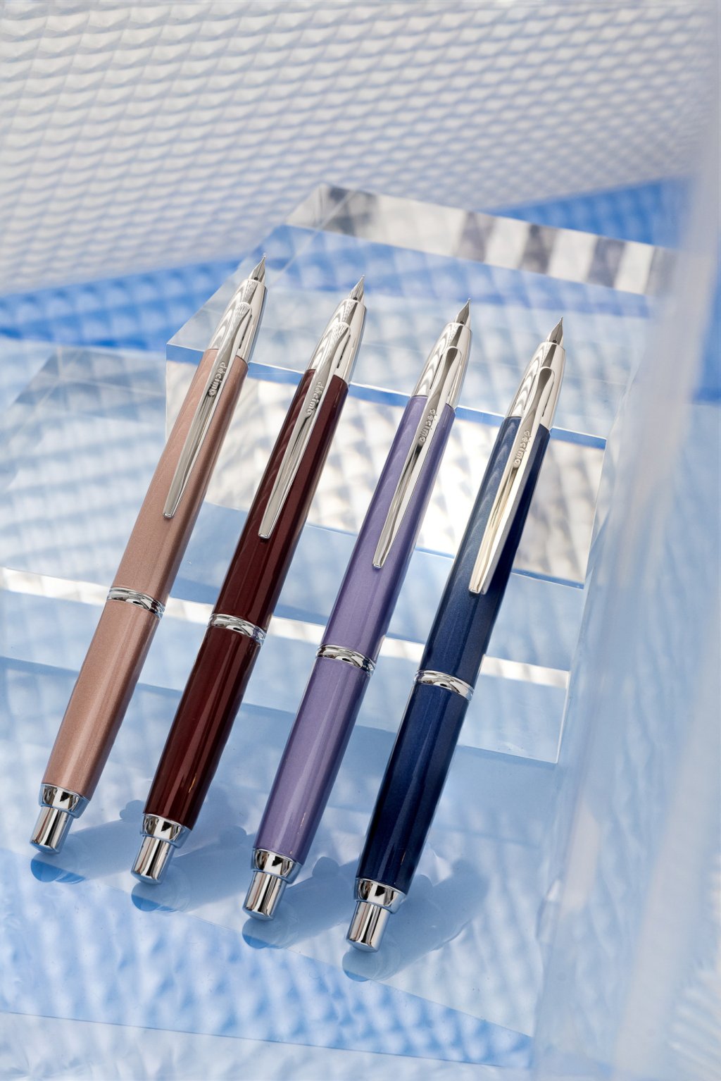 Pilot Capless Decimo Retractable Fountain Pens, also known as Vanishing Points, lined up on a clear block. There are four different coloured barrels: Champagne Pink, Red, Violet and Dark Blue.